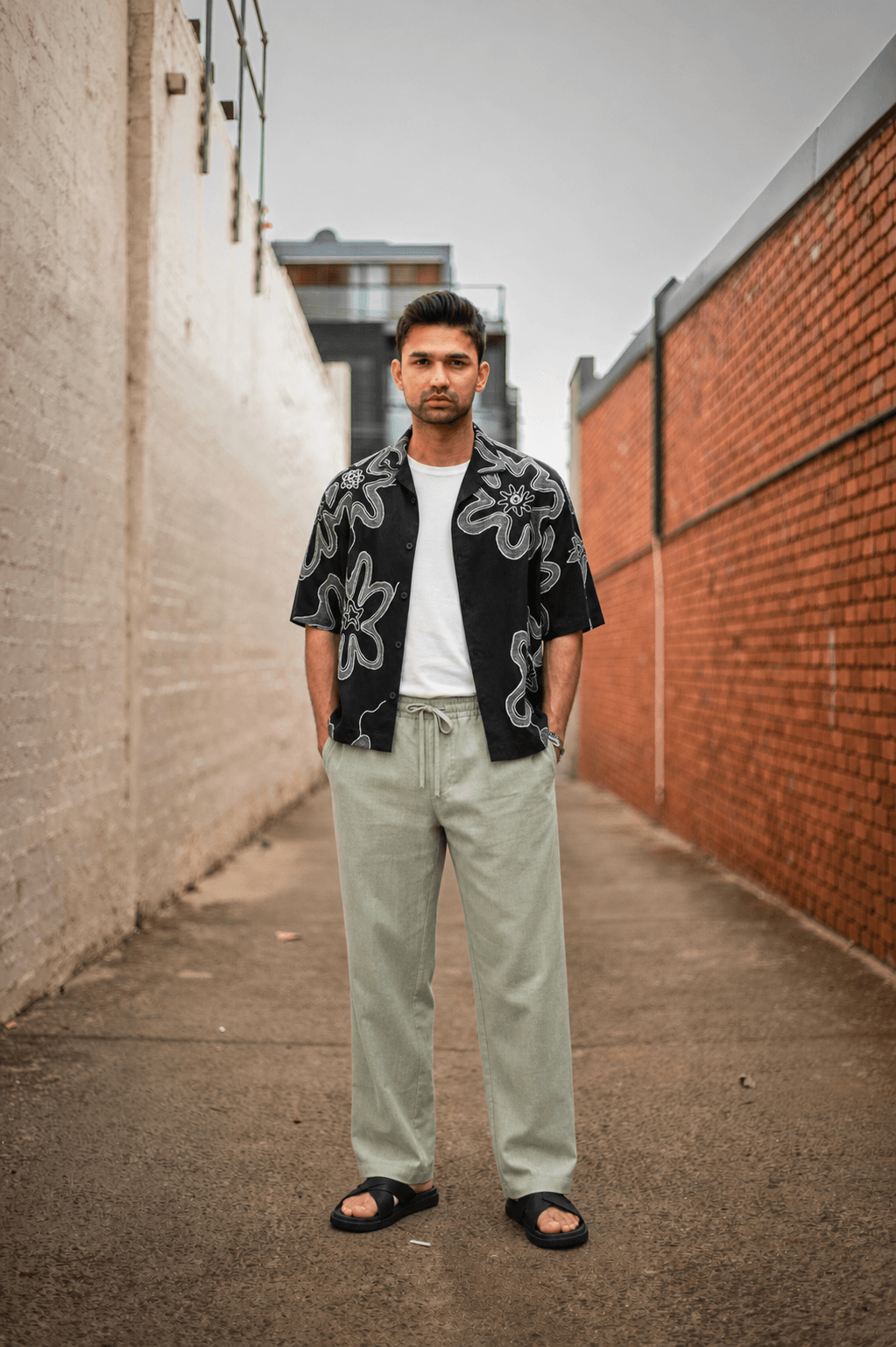 After: the same supplied real man model in the unchanged photo, wearing a black patterned short-sleeve shirt, white T-shirt, pale sage wide-leg trousers, and black sandals