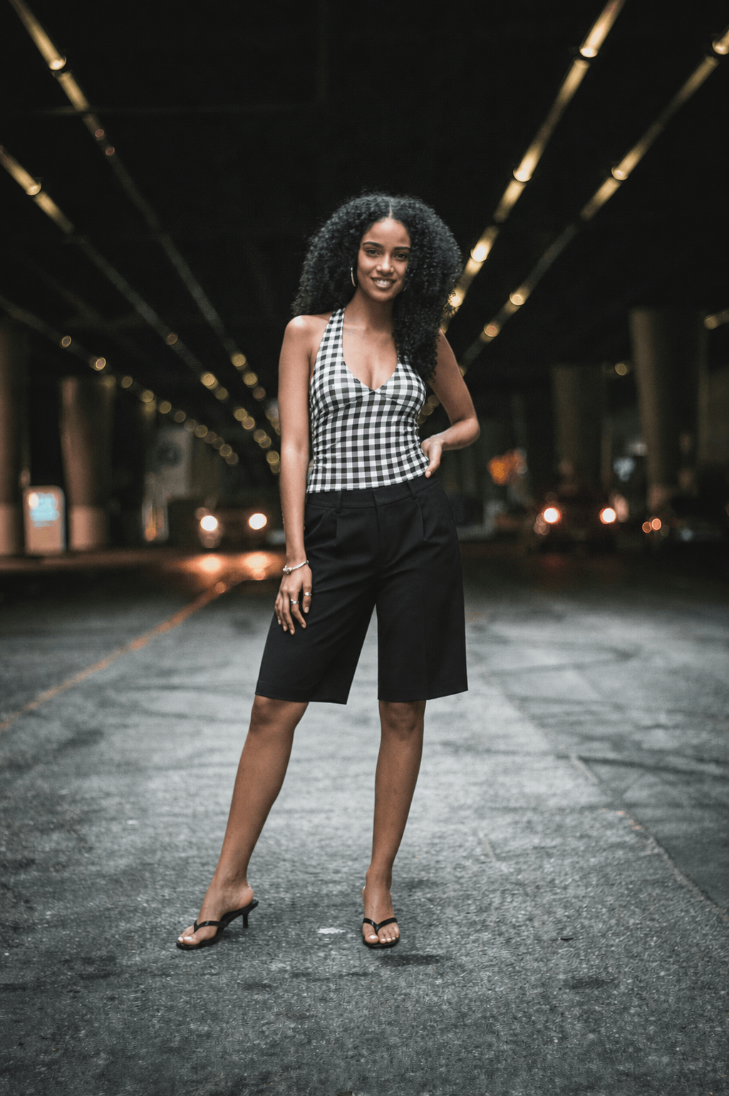The same supplied real woman model in the same photo, wearing the reference outfit: a gingham halterneck top, black tailored Bermuda shorts, and black kitten-heel mules
